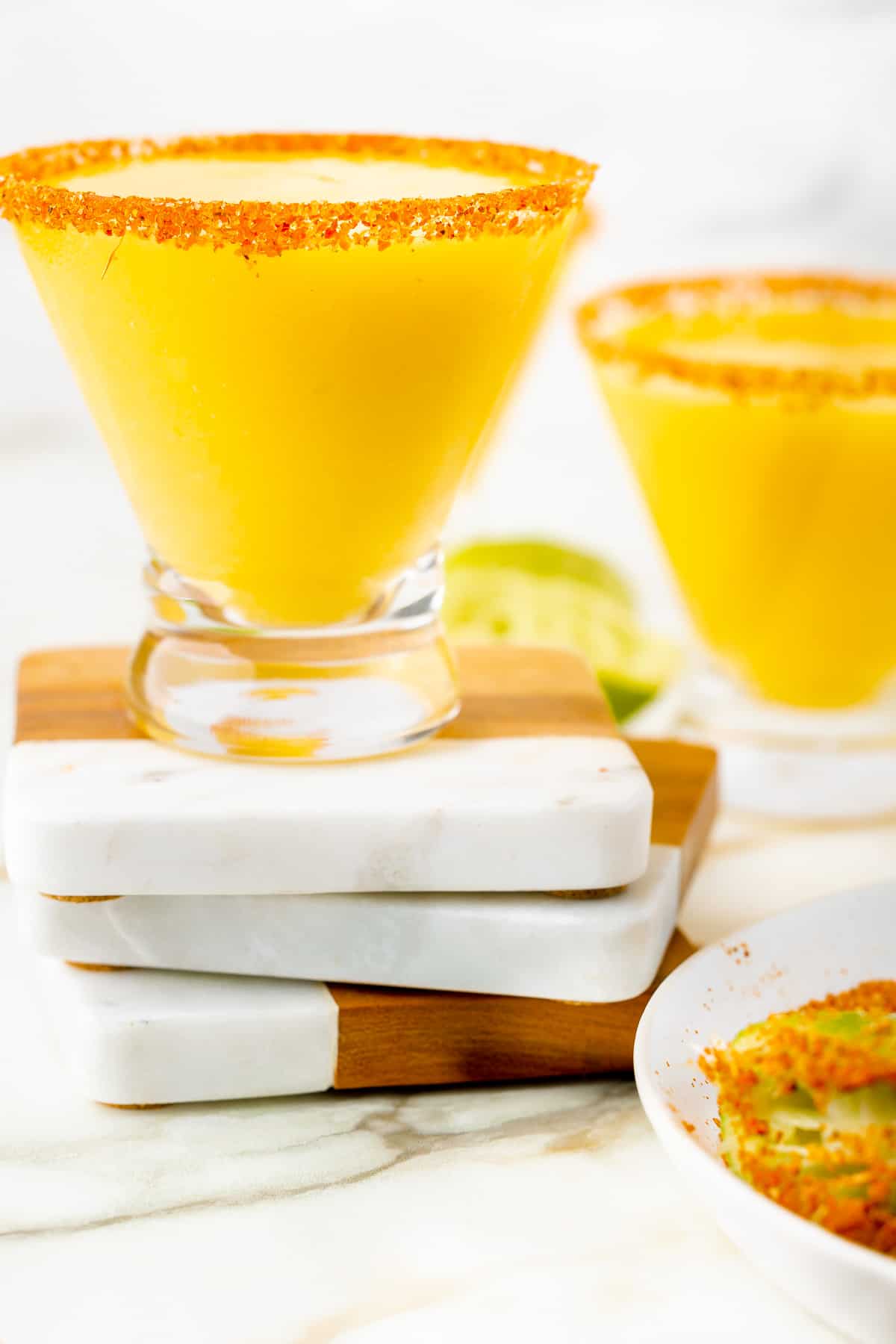 A yellow, frozen mangorita with red and orange seasoning on the rim, on top of three coasters