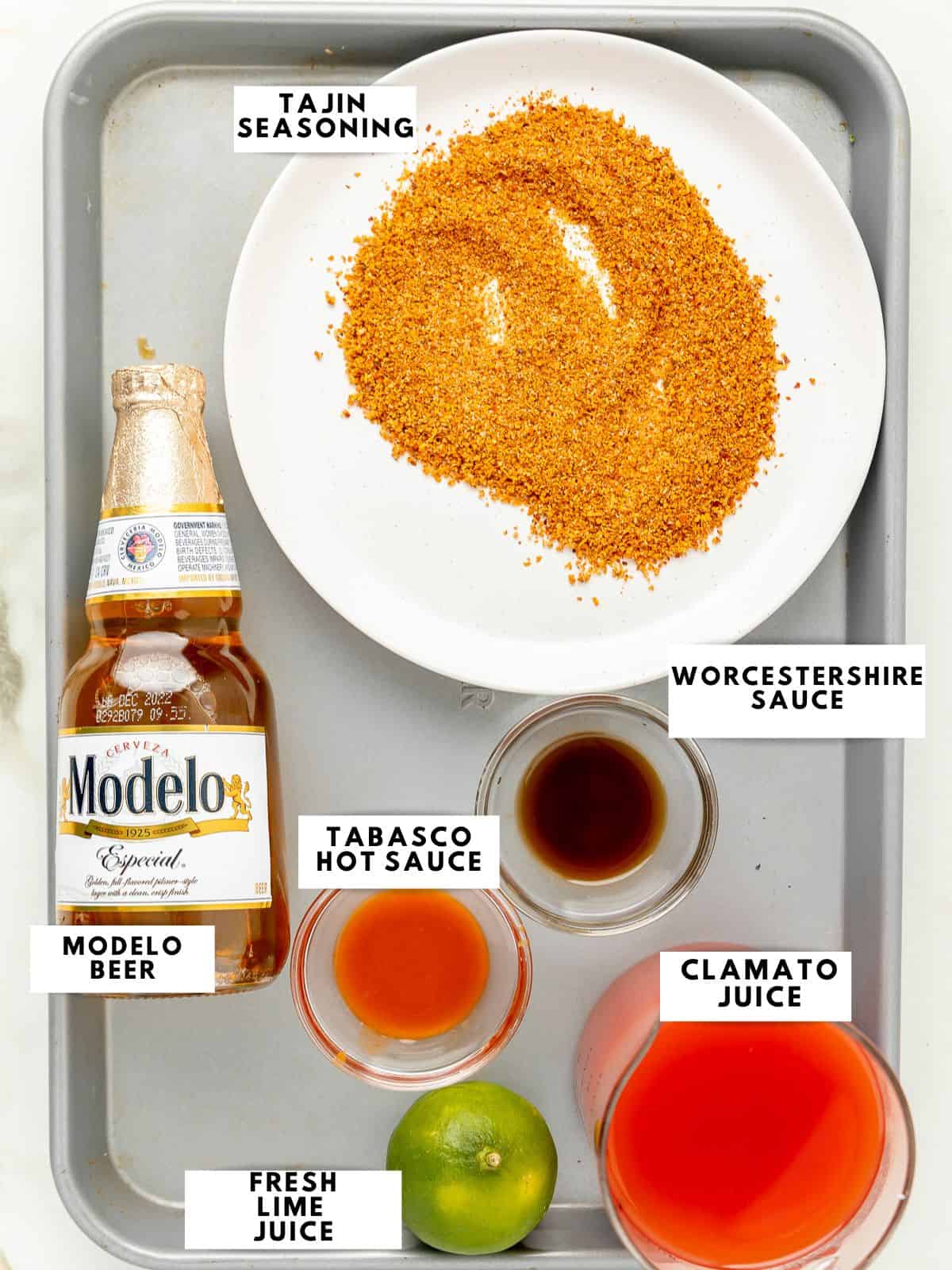 Photo of ingredients on a silver tray, tajin seasoning on. plate, a modelo beer, worcestershire sauce, tabasco hot sauce, clamato juice, fresh lime.