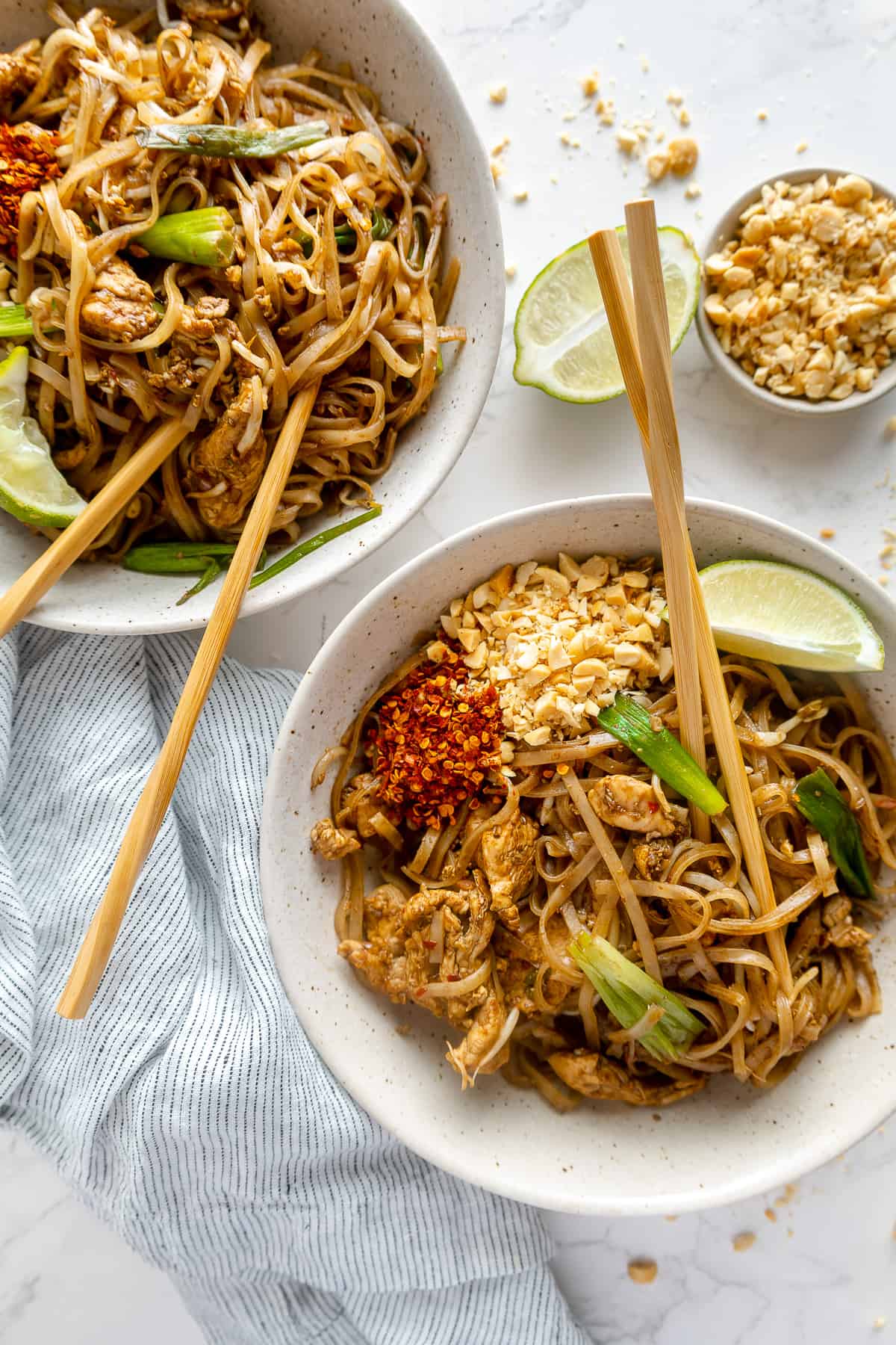 Two bowls of pad thai garnished with lime, peanuts and chilli flakes, with chopsticks. 
