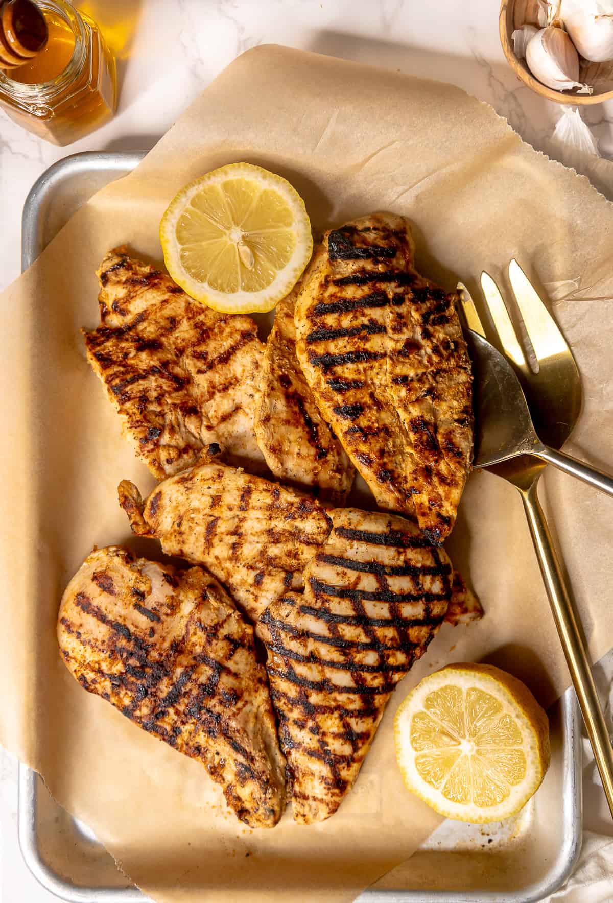 Grilled chicken breasts laid on a sheet pan, with two large serving forks and lemon to garnish.