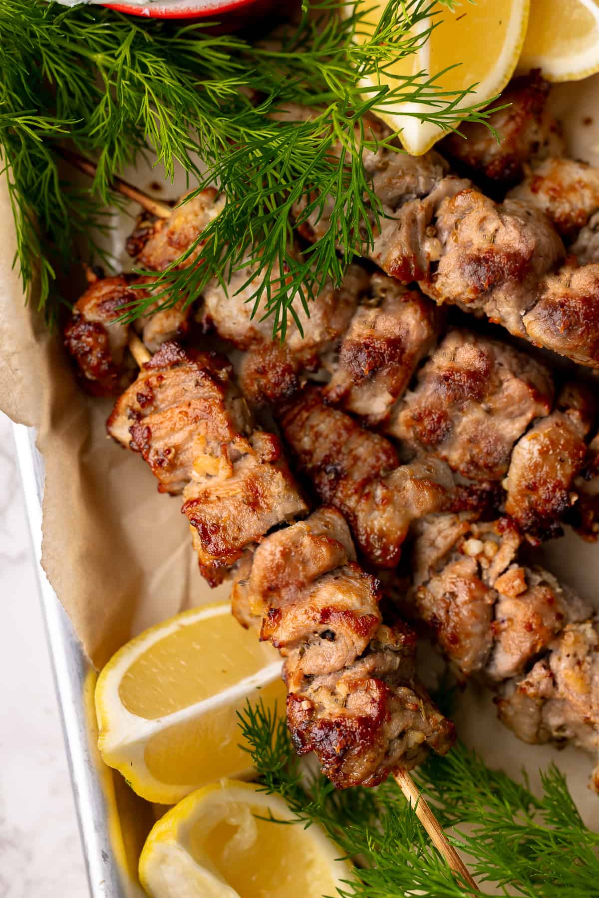 Cooked pork souvlaki served on a parchment-lined baking sheet, with lemon wedges and fresh dill. 
