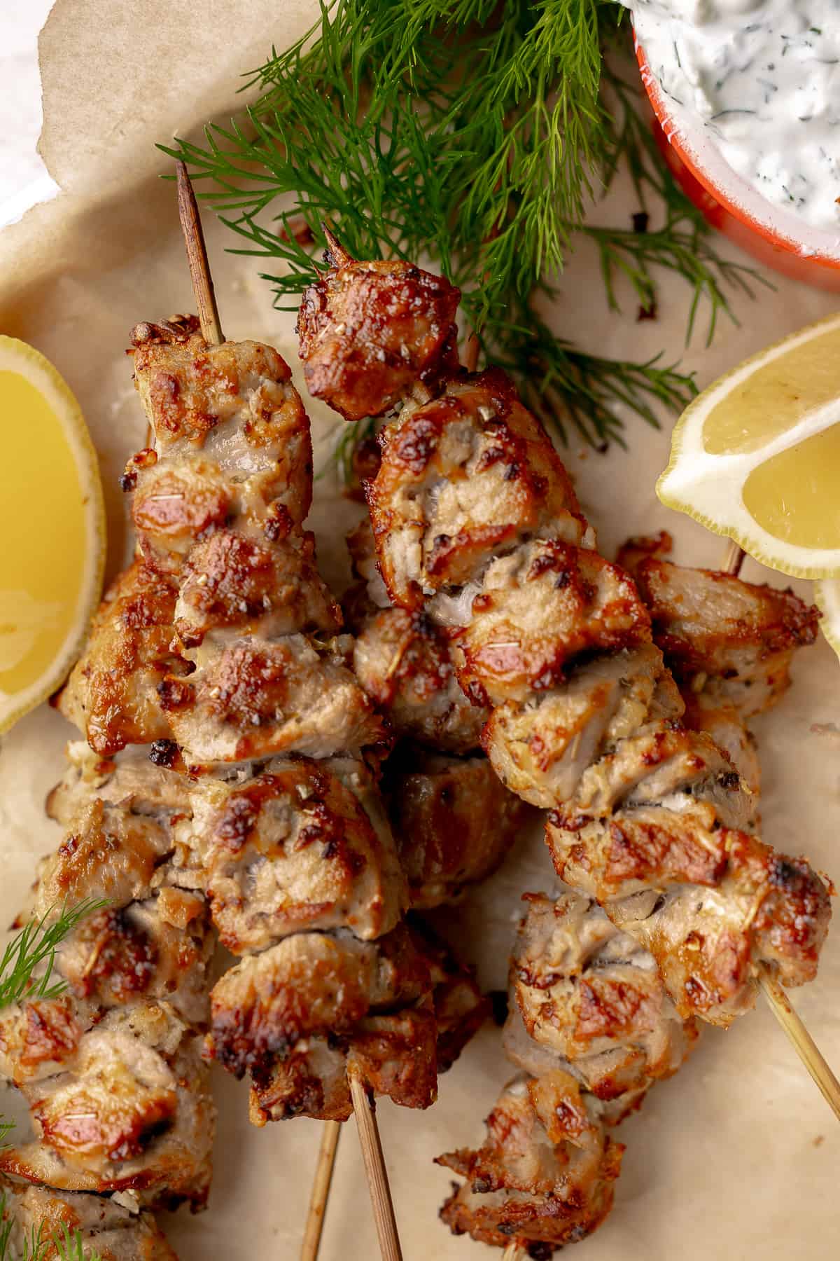 Skewers of pork souvlaki arranged on a sheet tray, with lemon and tzatziki. 