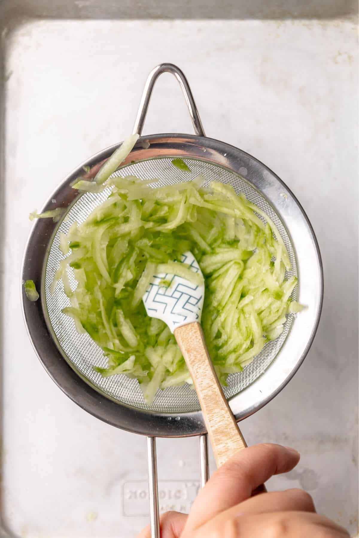 Shredded cucumber in a strainer, with a rubber spatula pressing liquid out.