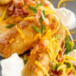 Mashed potato spring rolls served on a platter with cheddar cheese, green onions, and bacon on top.