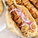 A chicken souvlaki pita with tzatziki and red onion, with chicken skewers in the background.